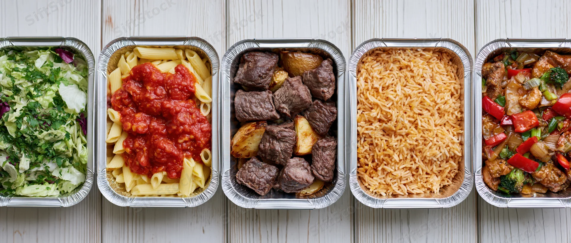Five aluminum takeout containers in a row each filled with different meals on a wooden surface