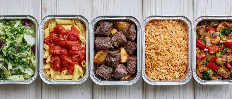 Five aluminum takeout containers in a row each filled with different meals on a wooden surface