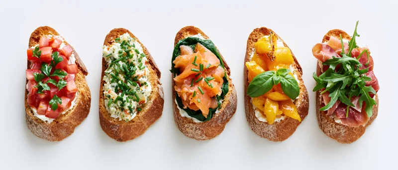 Five assorted bruschetta toasts arranged in a row on a white background