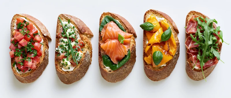Five assorted bruschetta toasts arranged in a row on a white surface