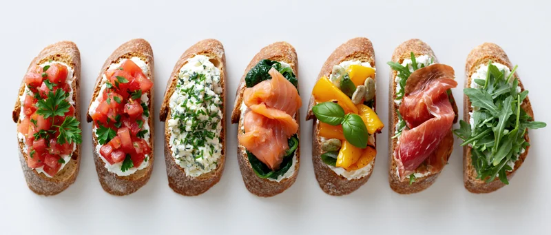 Seven bruschetta toasts with various toppings arranged in a row on a white surface
