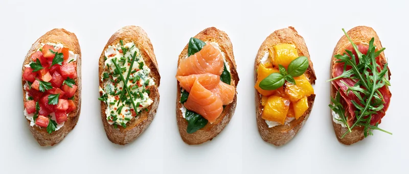 Five assorted bruschetta toasts arranged in a row on a white surface