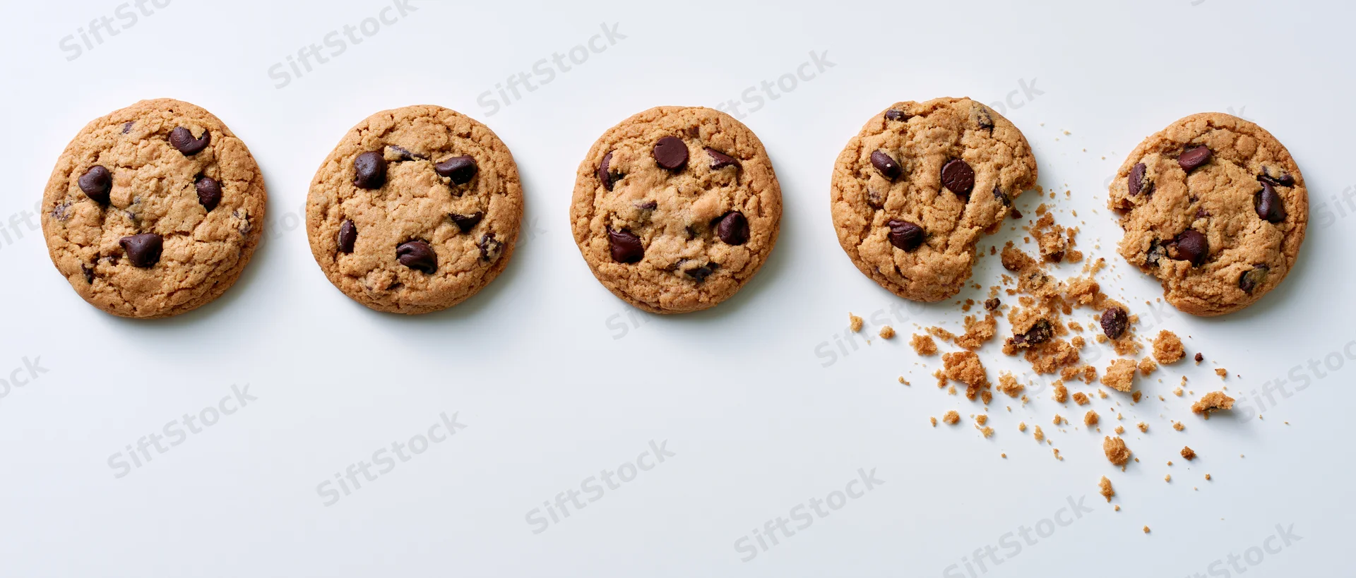 Five chocolate chip cookies in a row on white background with one broken and crumbs scattered