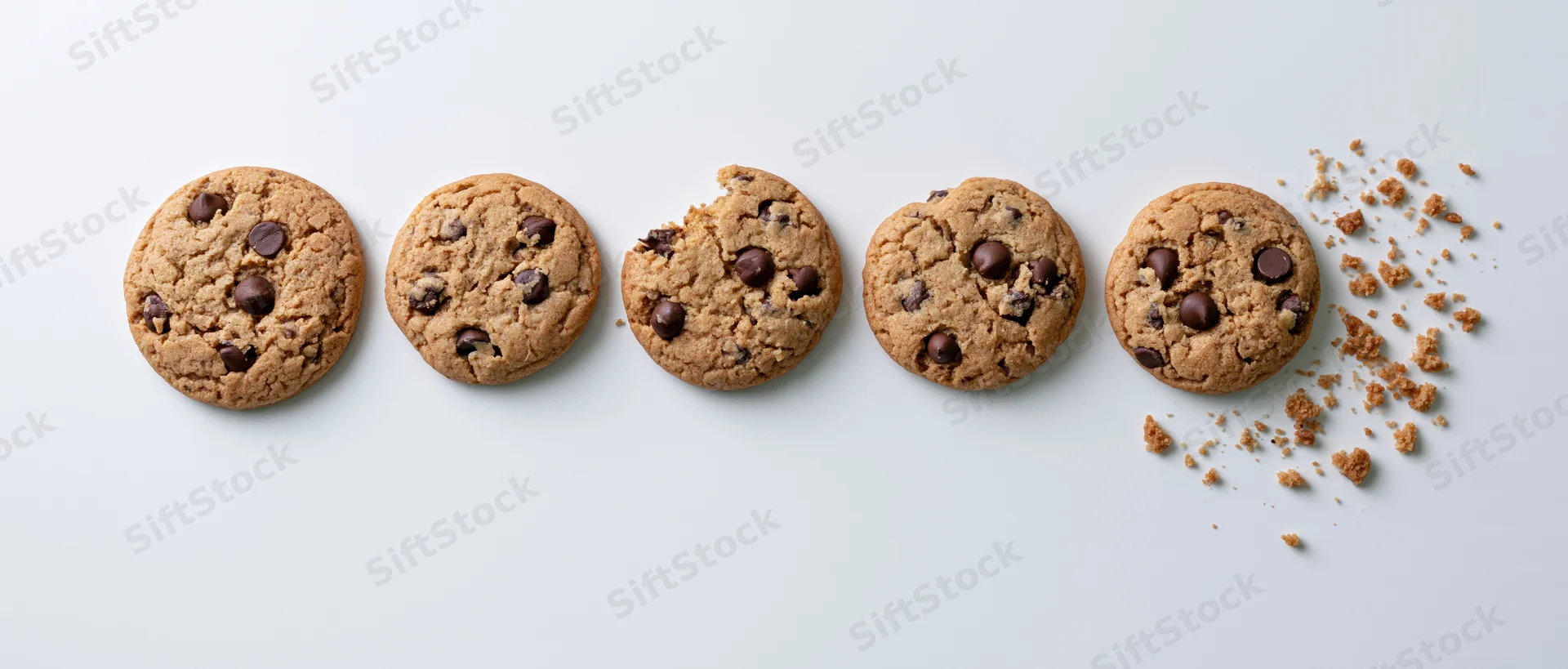 Five chocolate chip cookies in a horizontal row on a white surface, one partially eaten with crumbs scattered beside it