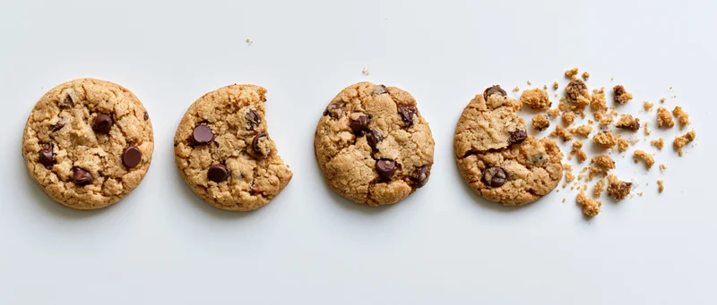 Four chocolate chip cookies in a row on white surface, one broken with crumbs scattered to the right