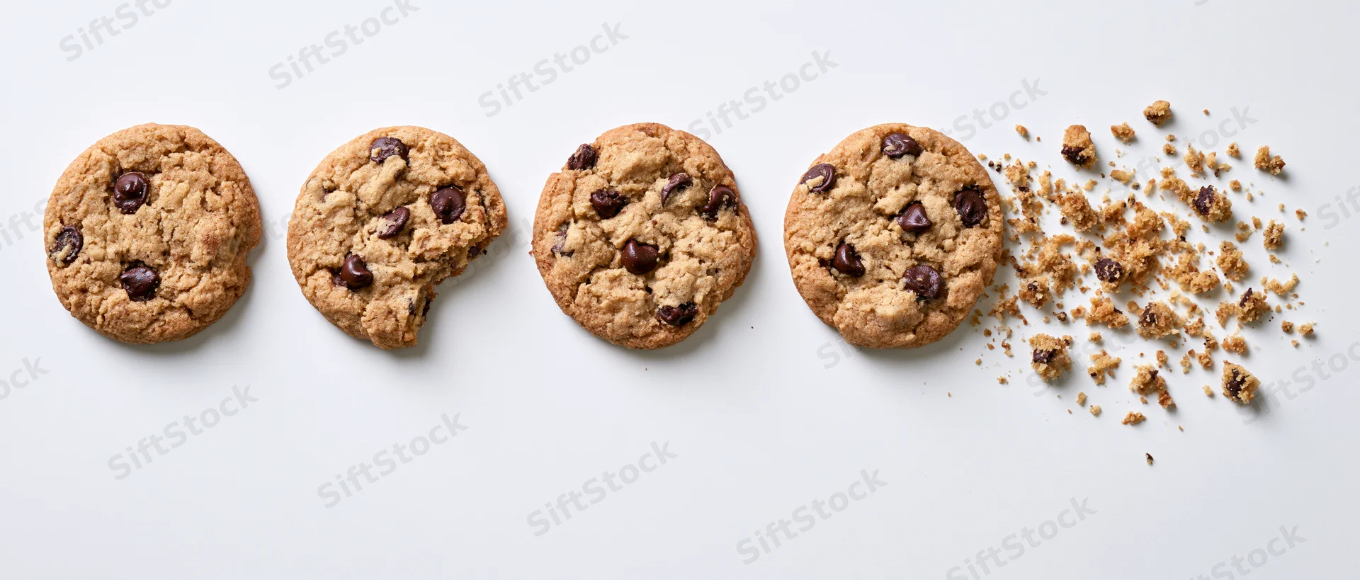 Four chocolate chip cookies in a row with one partially eaten and crumbs scattered on white background