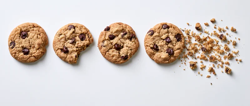 Four chocolate chip cookies in a row with one partially eaten and crumbs scattered on white background
