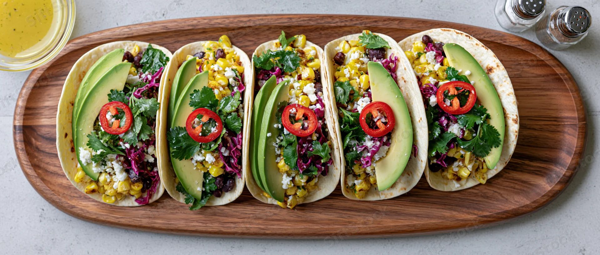 Five tacos with avocado, corn, black beans and cilantro arranged on a walnut serving platter