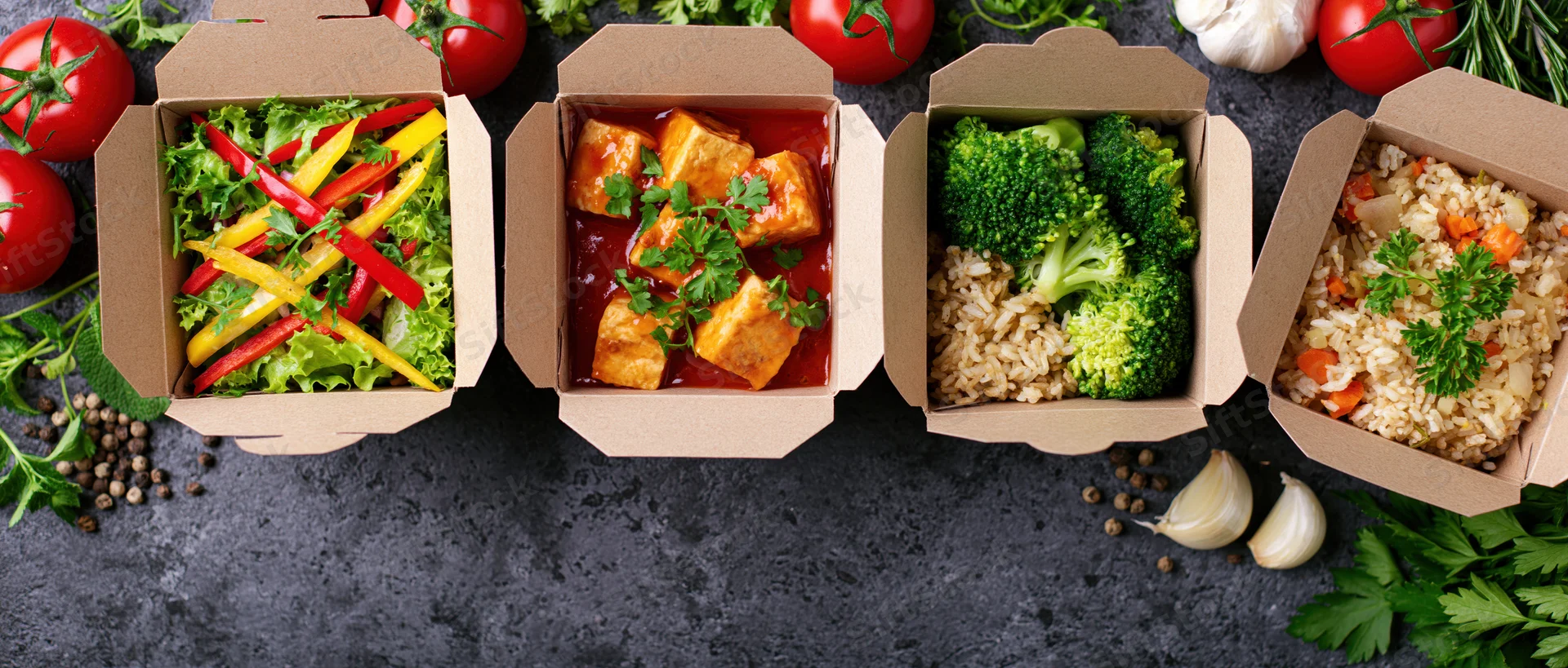 Four kraft paper takeout boxes in a row filled with different meals including salad, tofu in sauce, broccoli with rice, and fried rice, arranged on a dark countertop surrounded by fresh vegetables and herbs