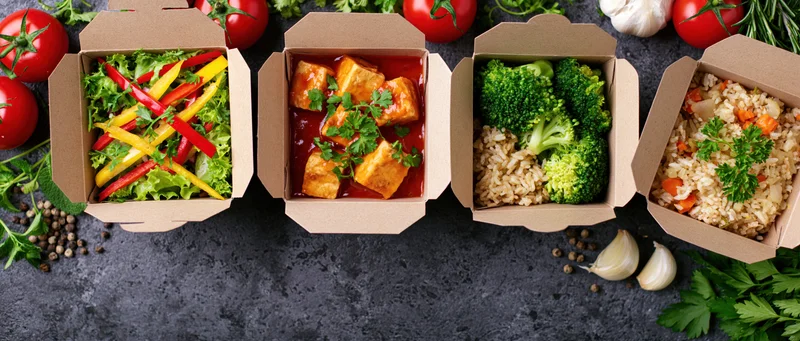 Four kraft paper takeout boxes in a row filled with different meals including salad, tofu in sauce, broccoli with rice, and fried rice, arranged on a dark countertop surrounded by fresh vegetables and herbs