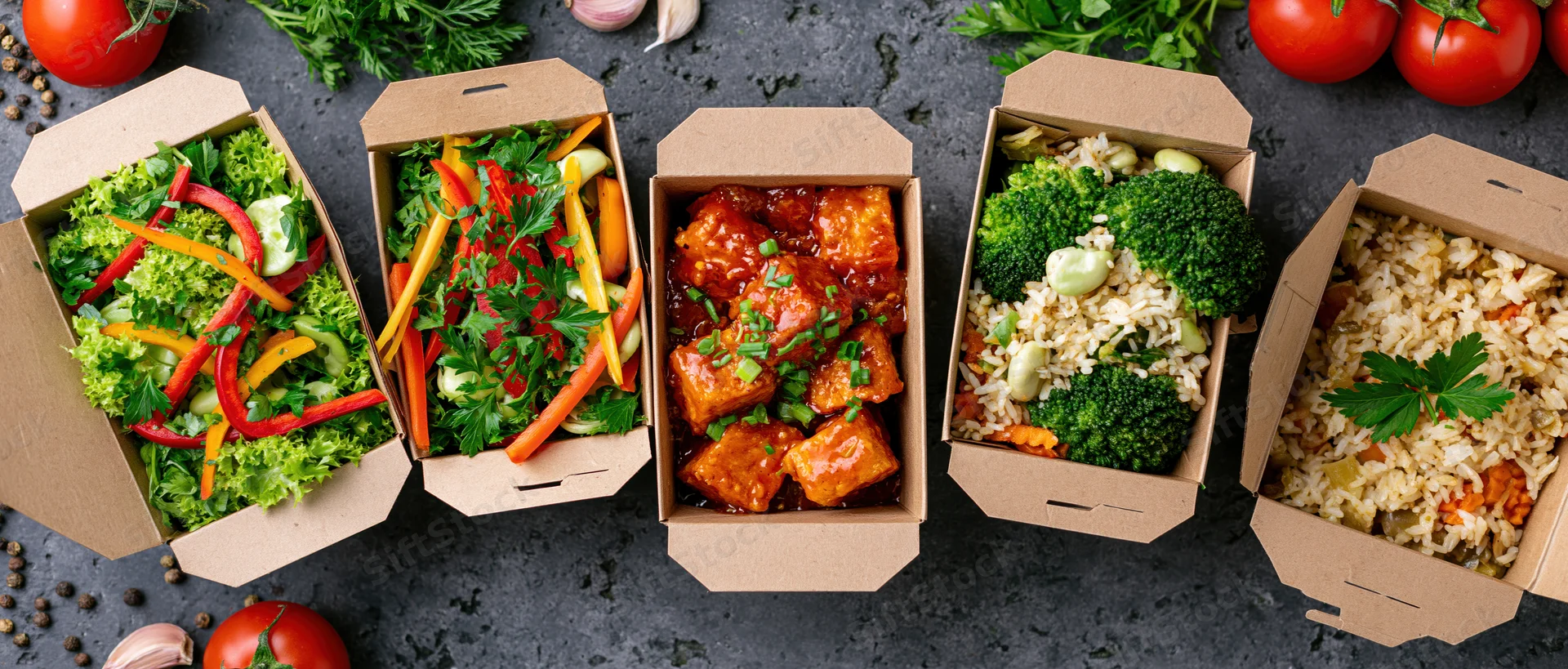 Five kraft paper takeout boxes arranged in a row filled with various meals including salads, tofu in sauce, and rice with vegetables on a dark surface