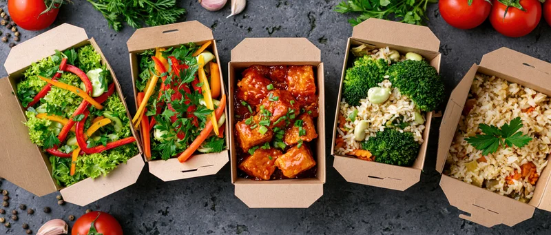Five kraft paper takeout boxes arranged in a row filled with various meals including salads, tofu in sauce, and rice with vegetables on a dark surface