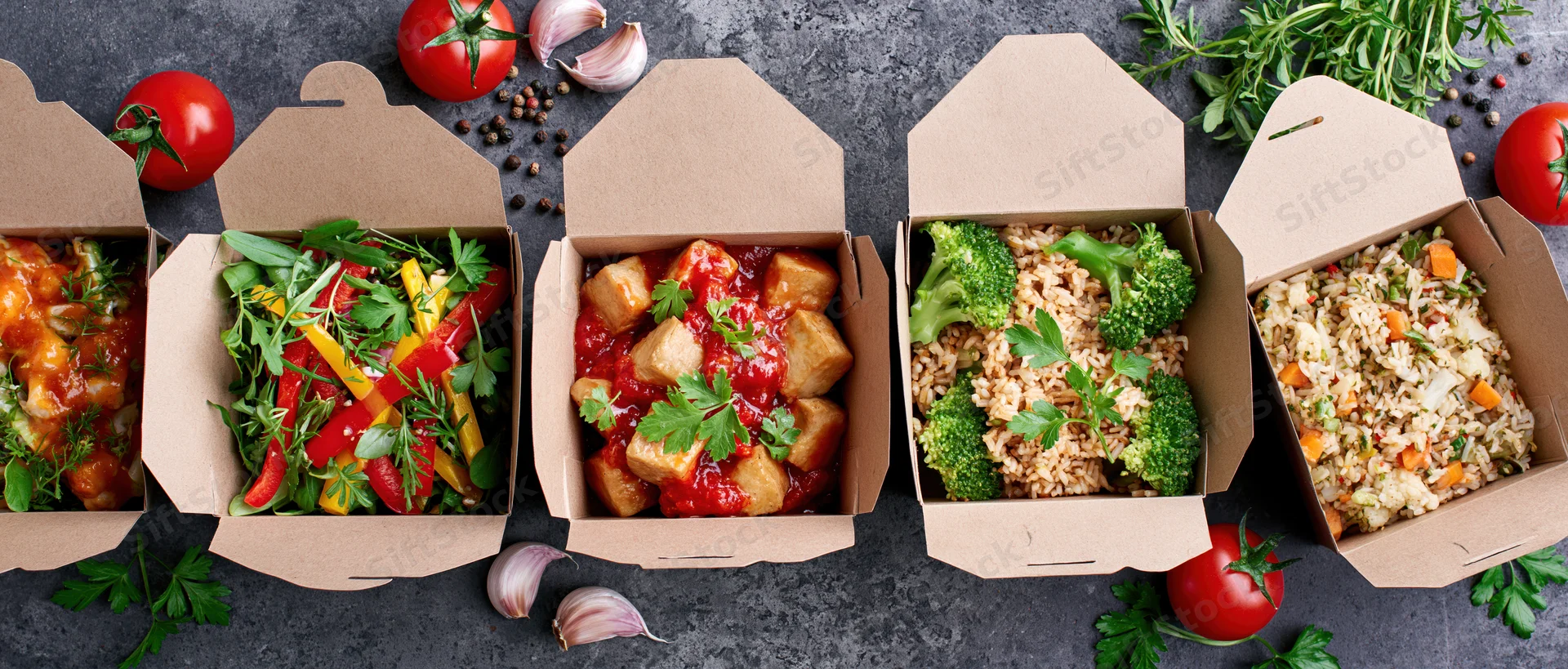 Five open kraft paper takeout boxes in a row filled with different meals on a dark surface surrounded by fresh herbs and vegetables