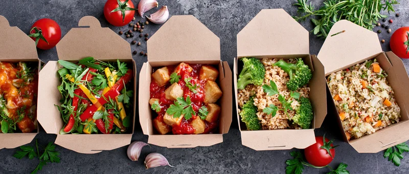 Five open kraft paper takeout boxes in a row filled with different meals on a dark surface surrounded by fresh herbs and vegetables