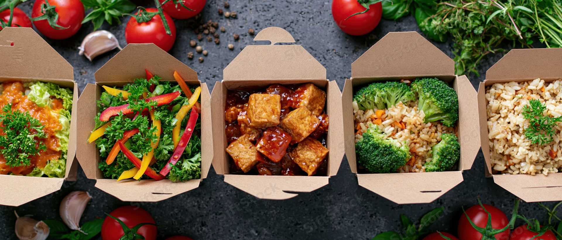 Five kraft paper takeout boxes in a row filled with different meals including tofu, broccoli, rice, salad, and vegetables on a dark surface surrounded by fresh herbs and tomatoes