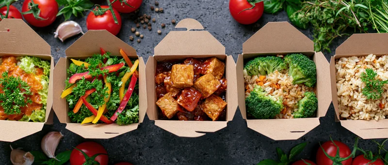 Five kraft paper takeout boxes in a row filled with different meals including tofu, broccoli, rice, salad, and vegetables on a dark surface surrounded by fresh herbs and tomatoes