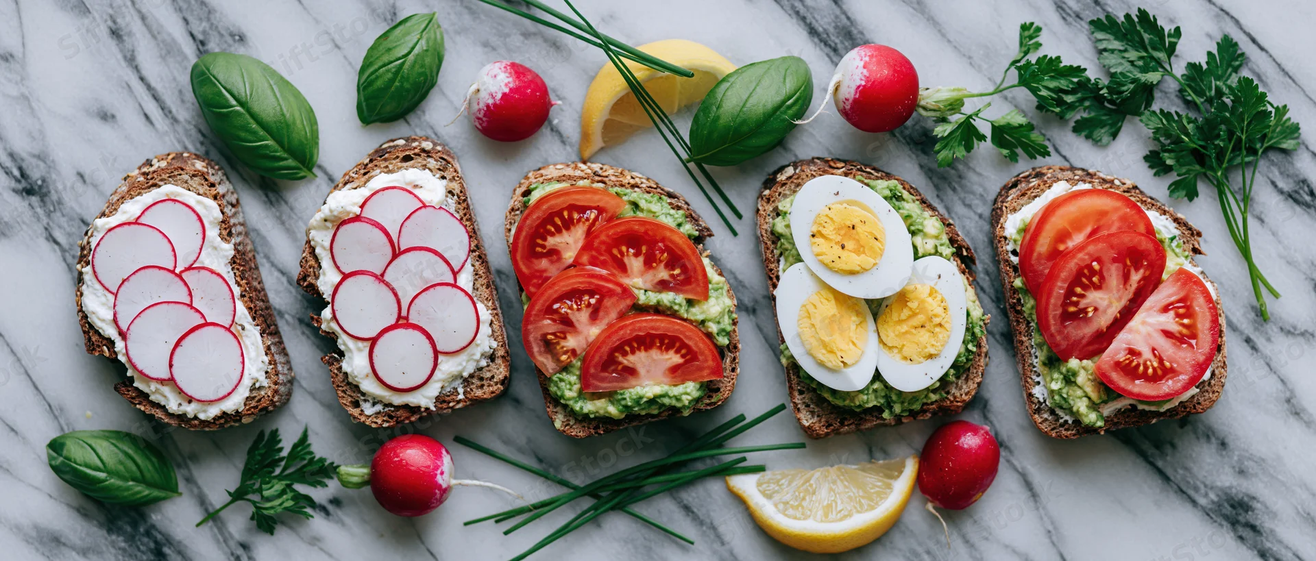Five openfaced sourdough toasts topped with avocado, tomato, radish, egg and herbs arranged in a row on a marble countertop