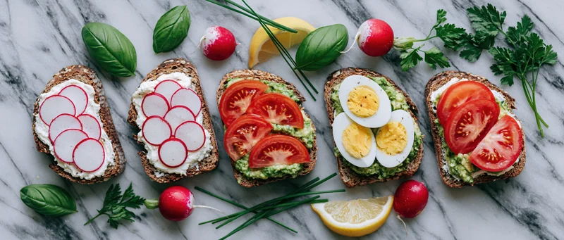 Five openfaced sourdough toasts topped with avocado, tomato, radish, egg and herbs arranged in a row on a marble countertop