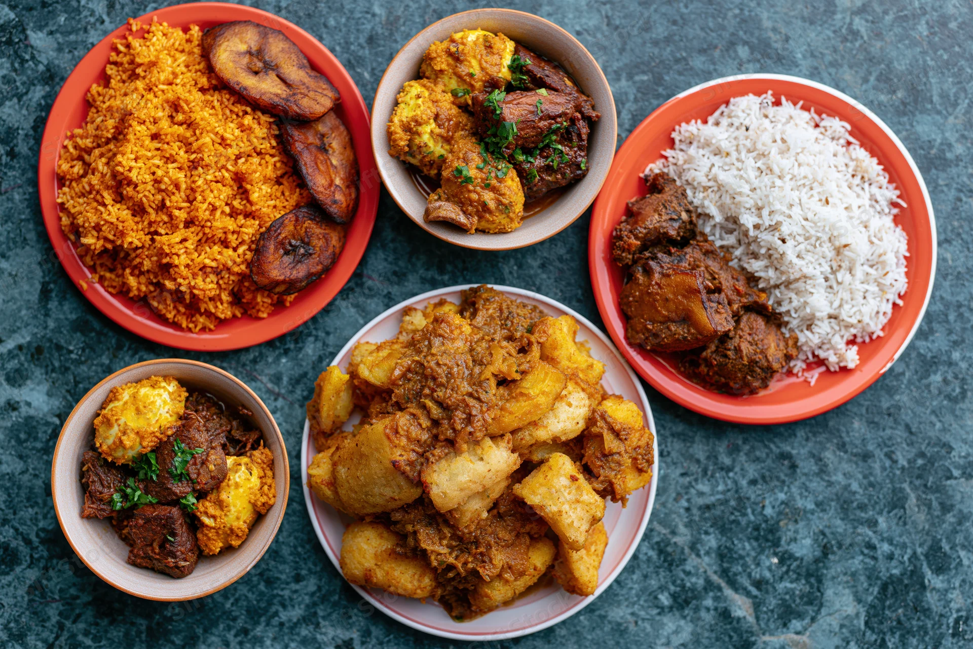 Five plates of west african food including jollof rice, stew, fried plantain and yam arranged on dark teal surface