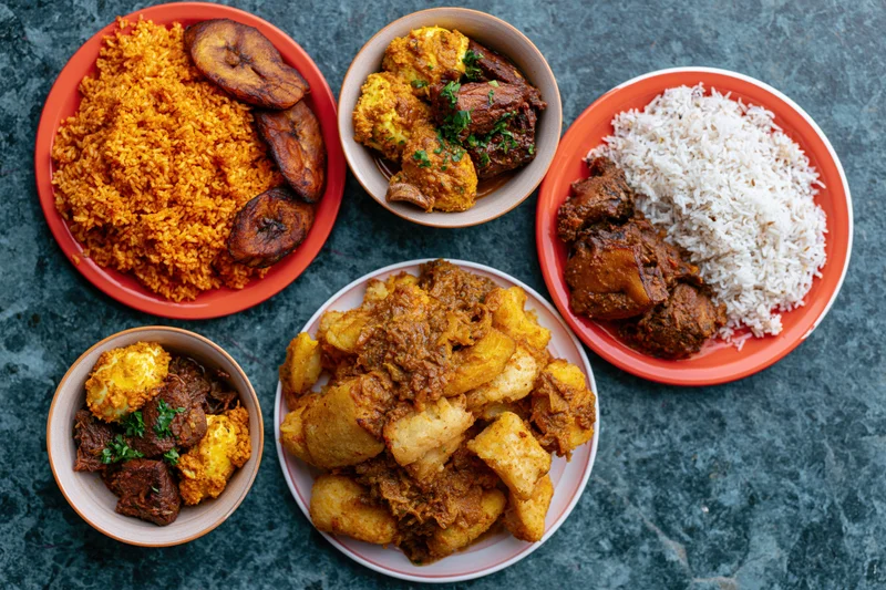 Five plates of west african food including jollof rice, stew, fried plantain and yam arranged on dark teal surface