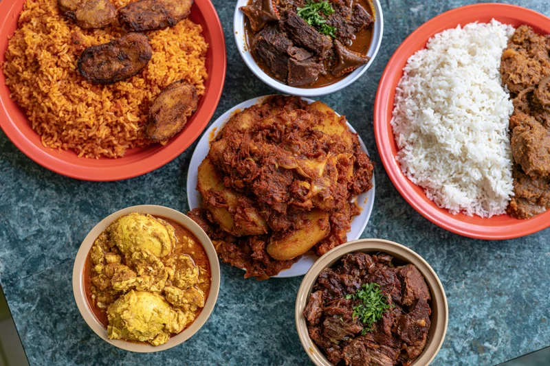 Several plates of West African dishes including rice, stews, plantains and yams arranged on a dark teal countertop