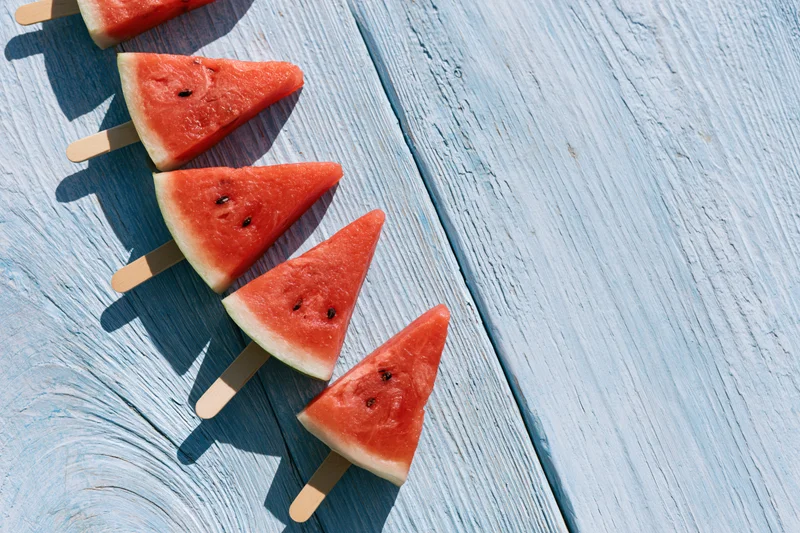 Triangular watermelon slices on wooden sticks arranged in a row on a blue wooden surface