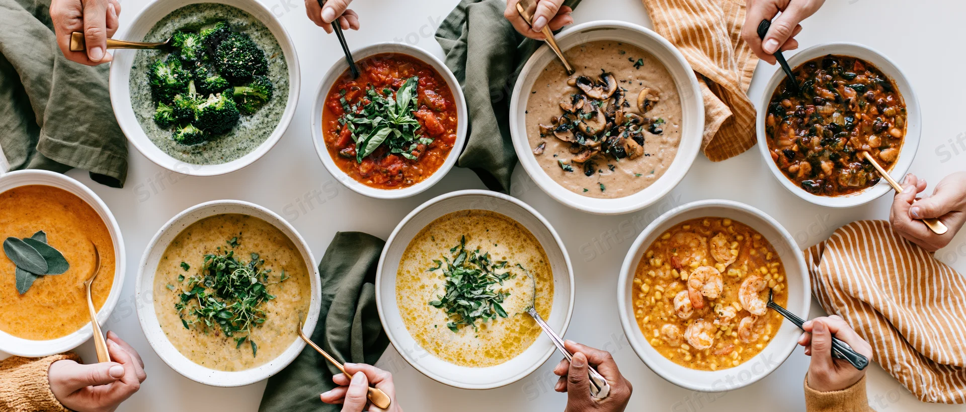 Eight bowls of varied soups and stews arranged overhead with hands holding spoons on a white table