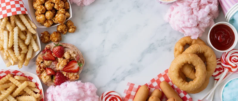 Carnival fair foods arranged in a border on a marble table with cotton candy, funnel cake, french fries, onion rings, and lollipops