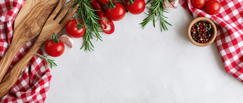 Fresh tomatoes, rosemary, garlic and wooden utensils arranged on a white countertop with red checkered cloth