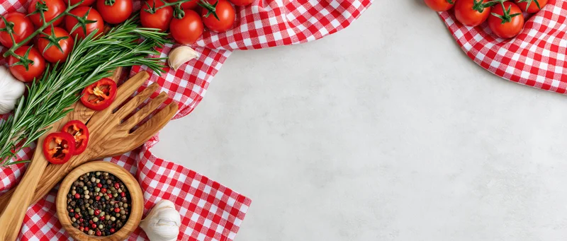 Fresh tomatoes, rosemary, garlic, and wooden utensils arranged on a red checkered cloth with copyspace on a white surface