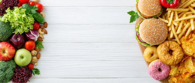 Healthy vegetables and fruits on one side, unhealthy fast food on the other, arranged on white wooden table in flat lay view with central copyspace