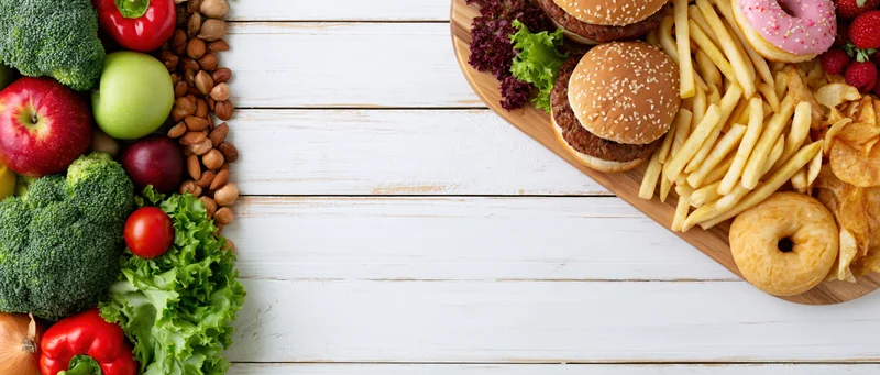 Healthy vegetables and fruits on one side, unhealthy fast food on the other, arranged on a white wooden table