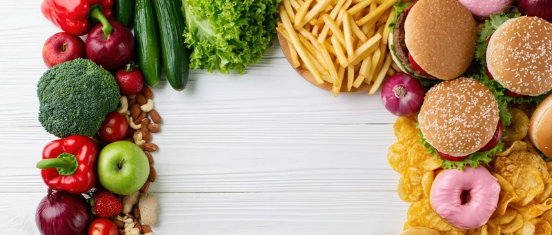 Healthy vegetables and fruits contrasted with unhealthy fast food on a white wooden table