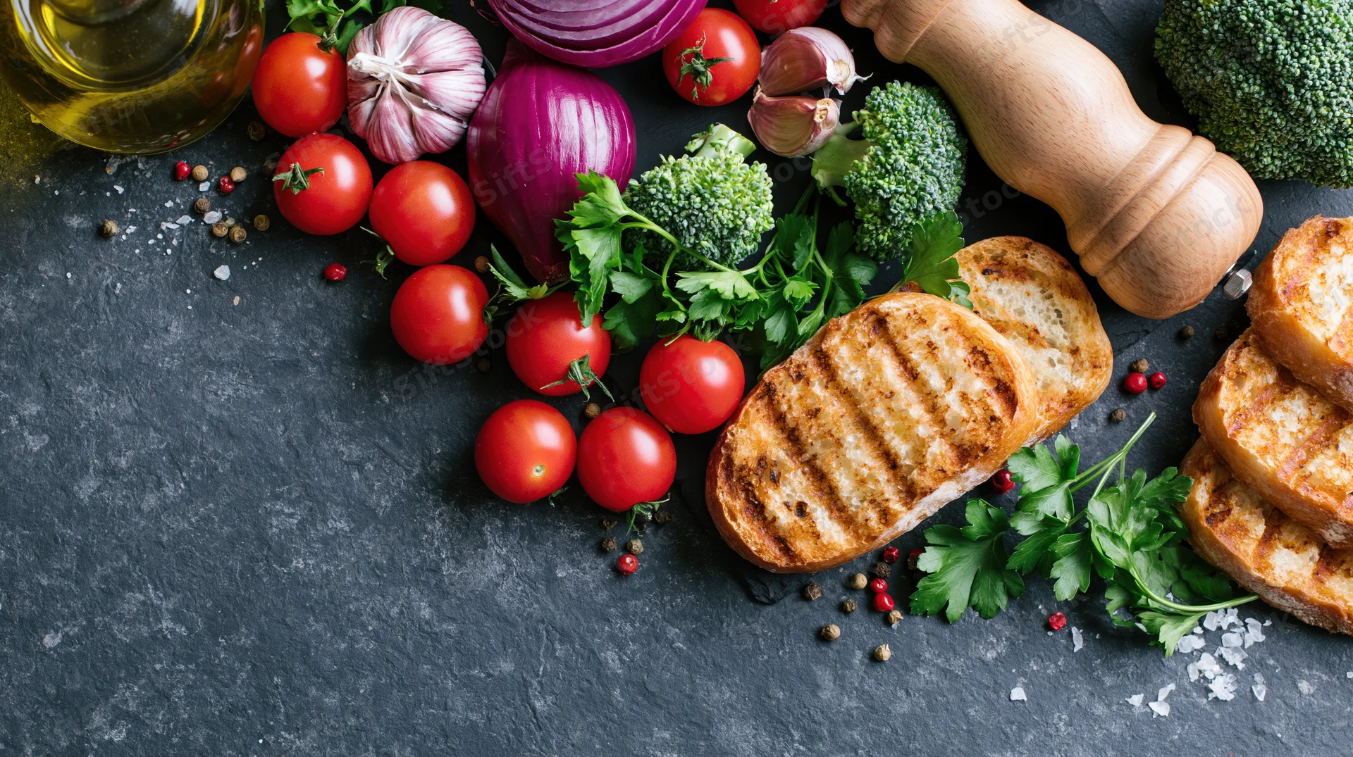 Fresh cooking ingredients arranged on dark slate surface with grilled bread and herbs