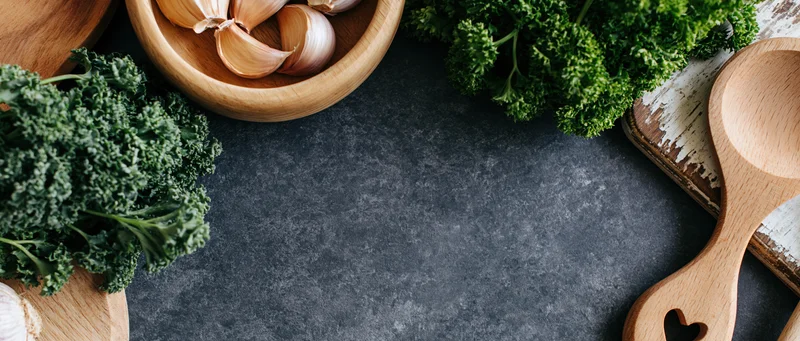 Fresh kale and parsley with garlic in wooden bowls and utensils arranged on dark countertop for flat lay food styling