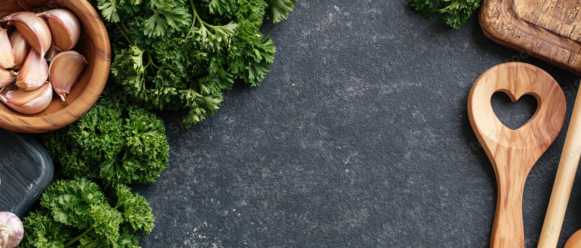 Fresh parsley and garlic in wooden bowl with heart-shaped spoon on dark stone countertop, flat lay arrangement