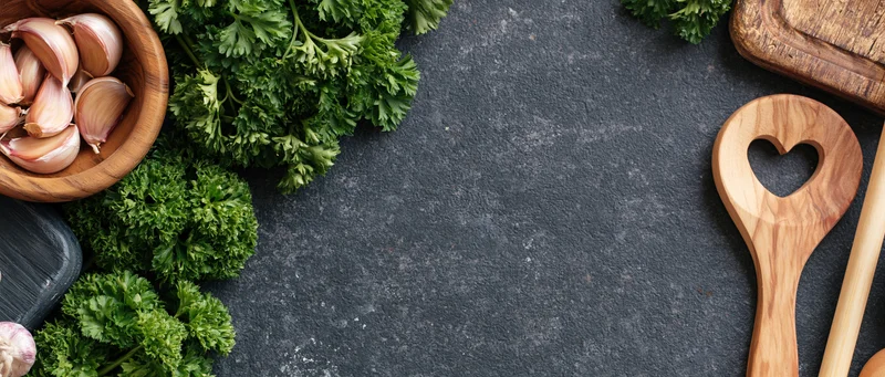Fresh parsley and garlic in wooden bowl with heart-shaped spoon on dark stone countertop, flat lay arrangement