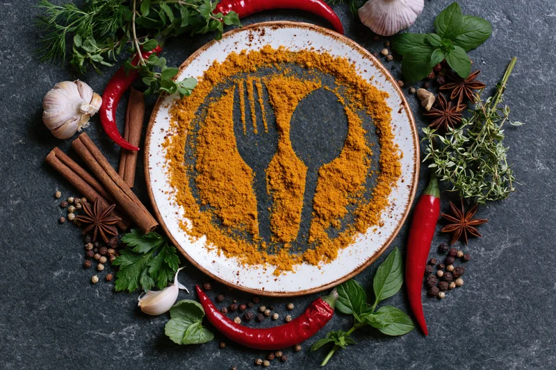Spice blend arranged on plate with fork and spoon shapes surrounded by fresh herbs and whole spices on dark slate surface