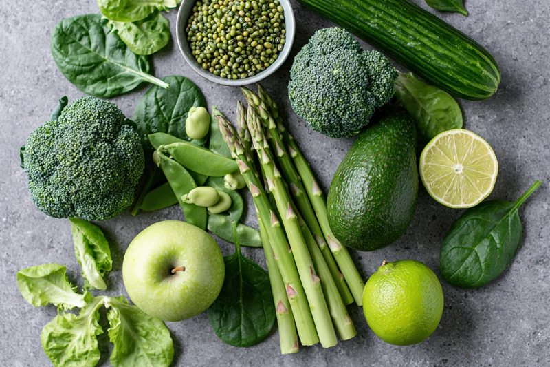 Assorted green vegetables and fruits arranged on a gray countertop in a flat lay composition