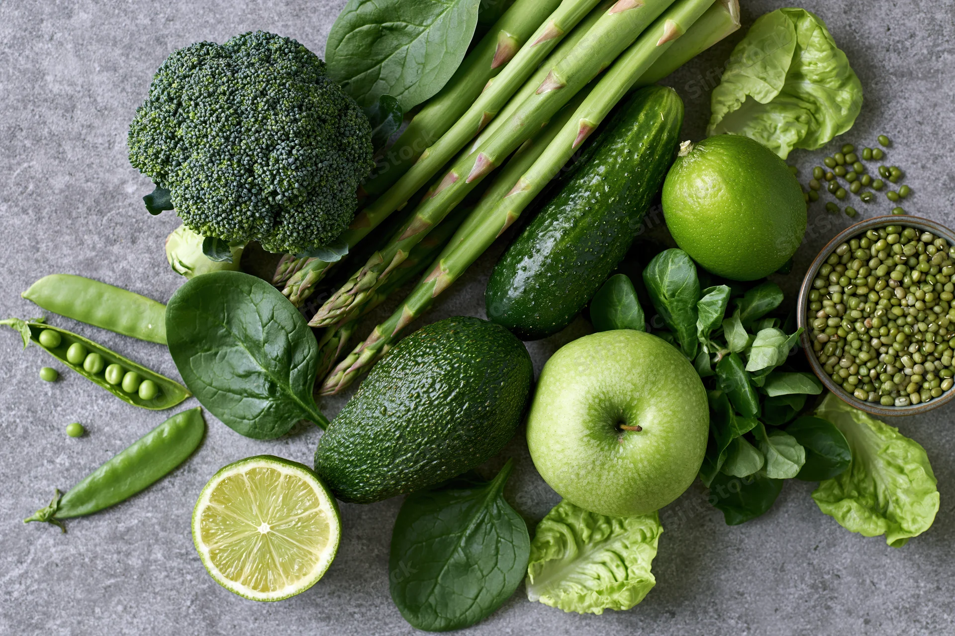 Assorted green vegetables and fruits arranged on a gray stone surface in a flat lay composition