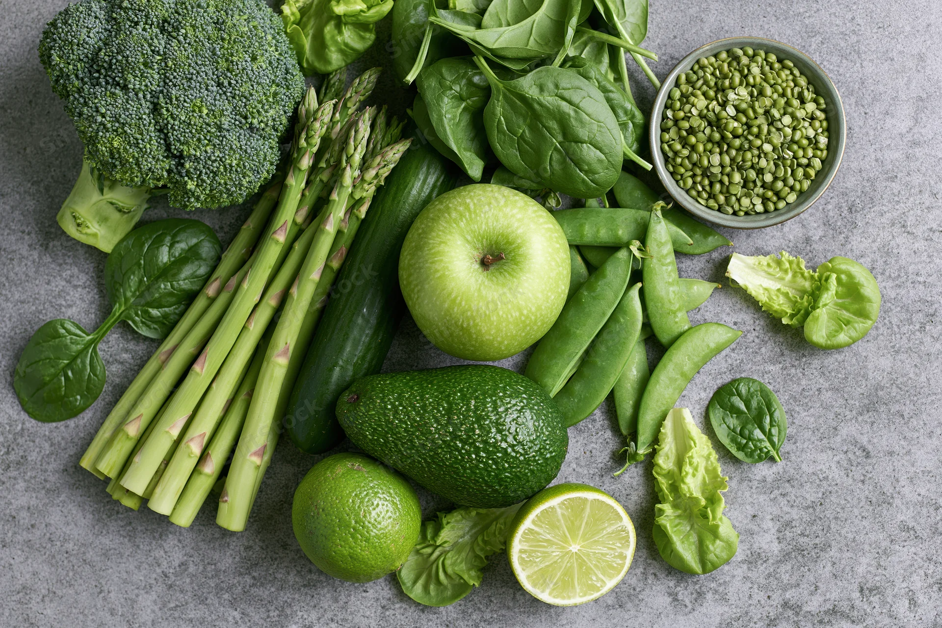 Assorted green vegetables and fruits arranged on a gray stone surface in a flat lay overhead view