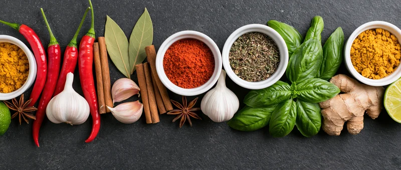Assorted spices and fresh herbs arranged in a row on a dark slate surface