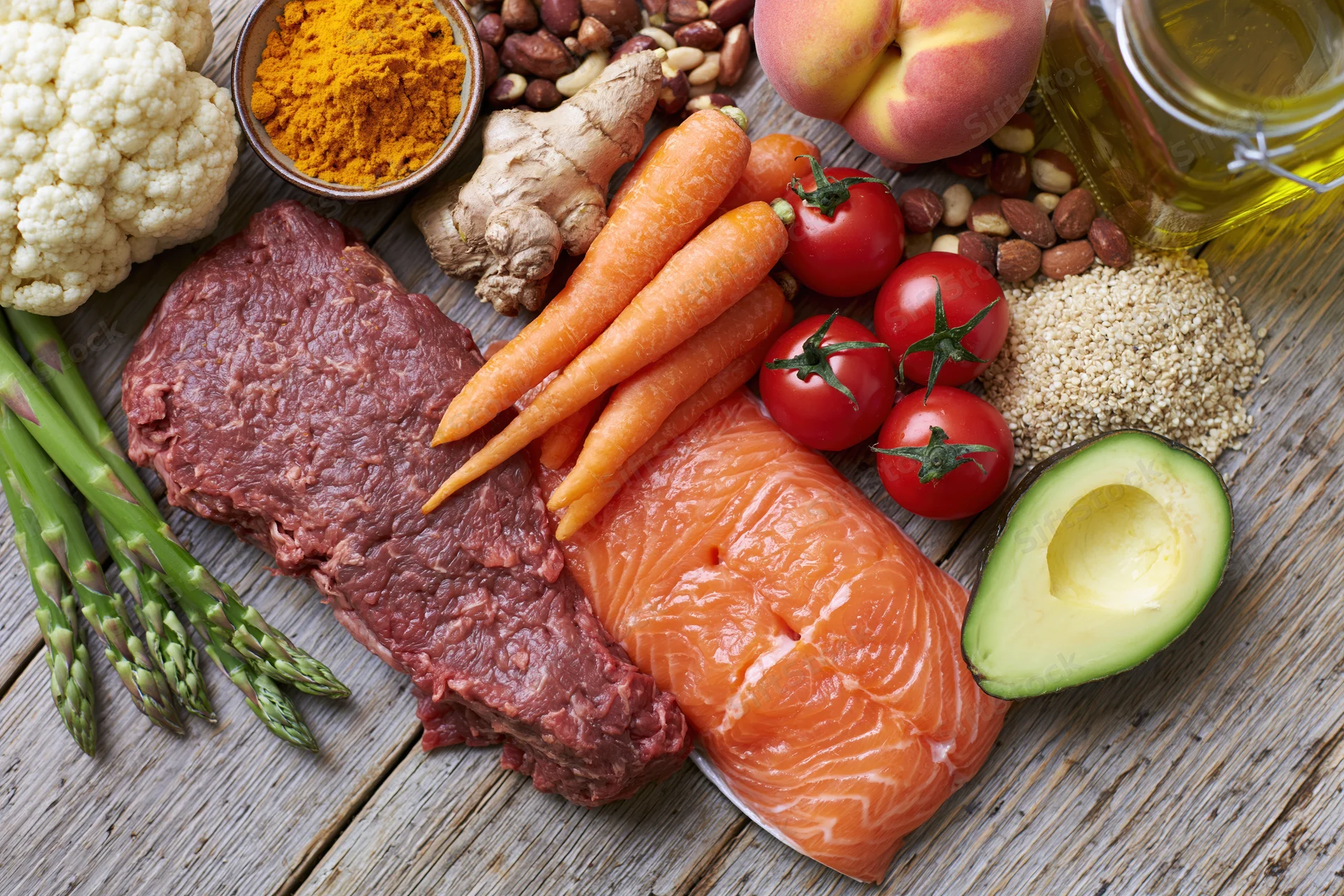 Variety of whole foods including meat, fish, vegetables, fruits, nuts and spices arranged on a wooden surface in a flat lay composition