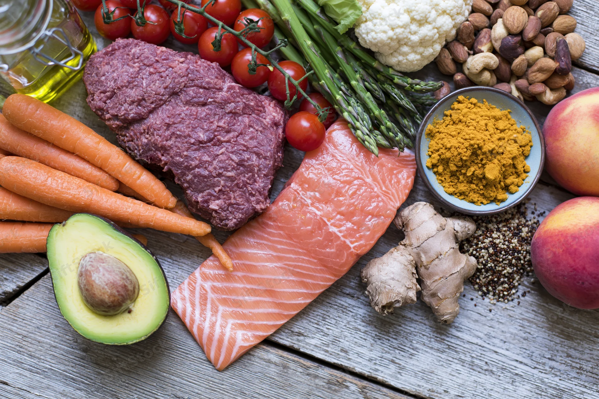 Salmon fillet, ground beef, avocado, carrots, tomatoes, asparagus, cauliflower, nuts, peaches, turmeric, ginger, quinoa, and olive oil arranged on a weathered wooden table