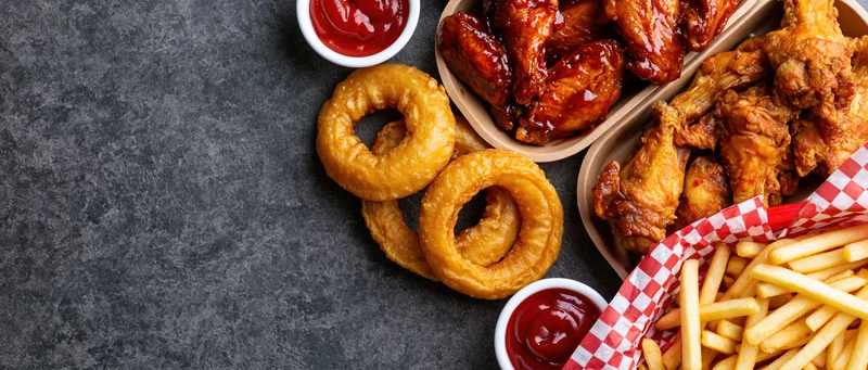 Bar food spread with chicken wings, onion rings, fries and ketchup arranged on dark surface with copyspace on left