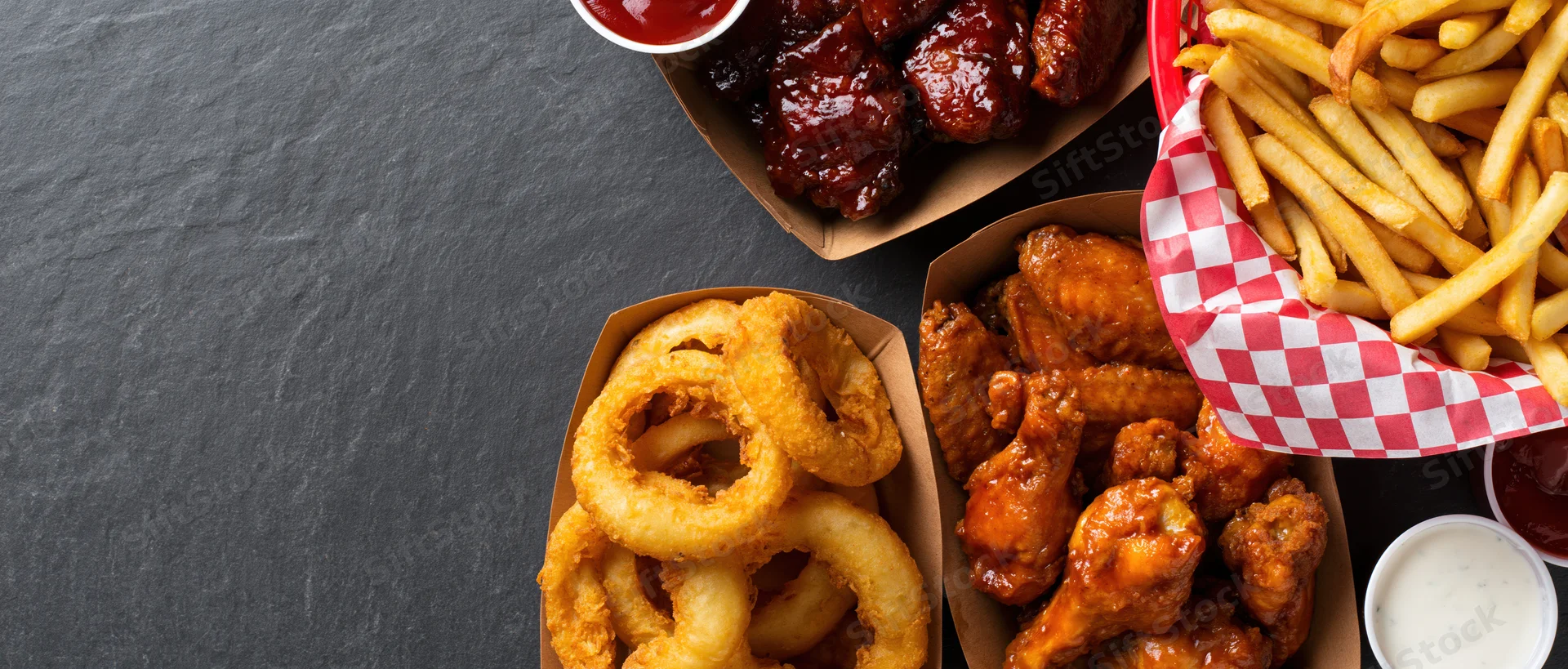 Bar food spread with wings, fries, onion rings and sauces arranged on dark surface with copyspace on left