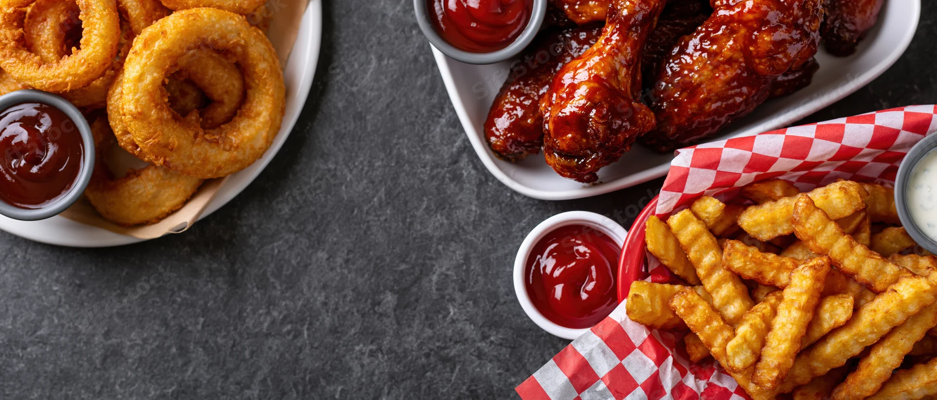 Bar food spread with onion rings, chicken wings and crinkle fries arranged on dark surface with dipping sauces and checkered paper