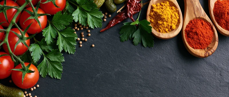 Cherry tomatoes on the vine with wooden spoons holding spices and herbs arranged on a dark slate surface