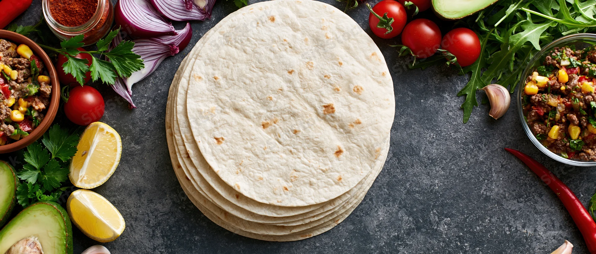 Stack of flour tortillas surrounded by fresh vegetables and salsa on dark textured surface