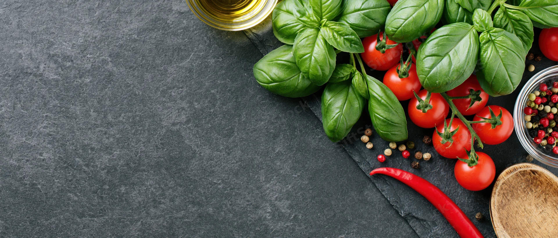 Fresh basil and cherry tomatoes on vine arranged with spices and oil on dark slate surface